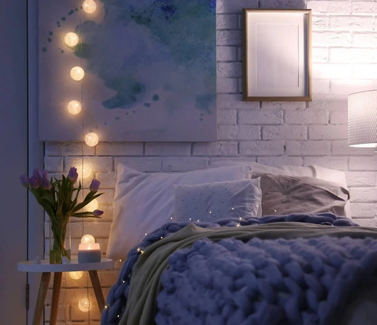 The bed with a purple quilt has warm-colored string lights above a side vase for mood lighting.